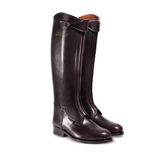 Puede incluir: Botas de montar de cuero negro con cremallera en la parte posterior y una correa de hebilla en la parte superior. Las botas tienen una suela de cuero marrón y un tacón bajo.