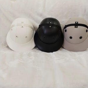 Könnte beinhalten: Drei Tropenhelme in einer Reihe, bestehend aus einem weißen Helm links, einem schwarzen Helm in der Mitte und einem beigen Helm rechts. Jeder Helm hat eine abgerundete Krone, eine breite Krempe, seitliche Belüftungsösen und einen Riemen.