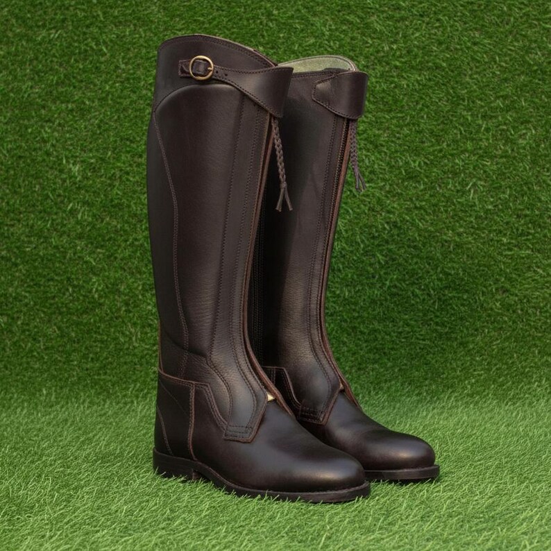 May include: A pair of tall, dark brown leather riding boots featuring a structured shaft, a rounded toe, and a side zipper closure. Each boot includes a decorative strap with a brass-toned metal buckle at the top and a braided leather pull tab.