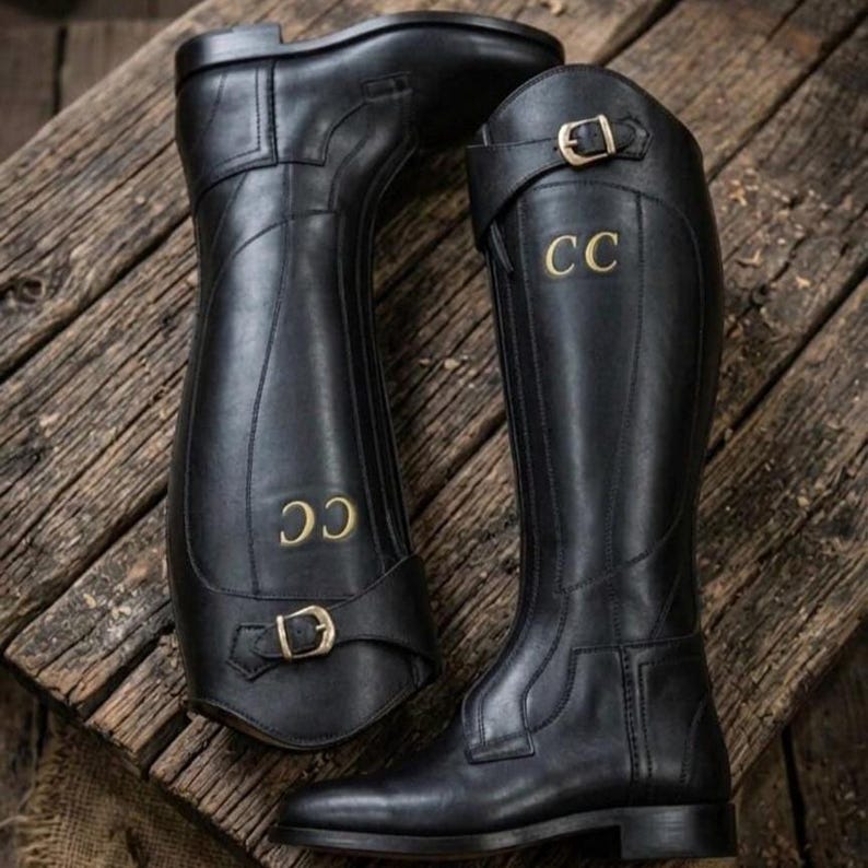 May include: A pair of black knee-high riding boots featuring a smooth finish, a low block heel, and a decorative strap with a gold-toned metal buckle at the ankle. The letters CC are embossed in gold on the outer side of each boot shaft.