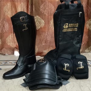 May include: A black tall riding boot with a buckle strap, a black protective knee guard, two black padded knee pads, and a black equipment bag. The bag features the gold text BROAD POLO WEAR, WWW.BWMFG.COM, and @BROADPOLOWEAR. All items feature a gold logo.