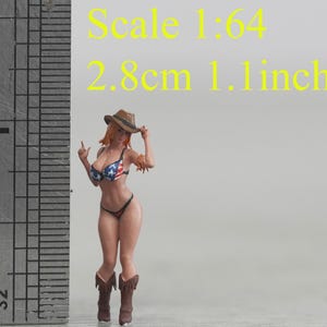 May include: Miniature figurine of a woman in a bikini with an American flag design, cowboy boots, and a hat. The figure is approximately 1.1 inches tall. The text "Scale 1:64" and "2.8cm 1.1 inch" is visible.