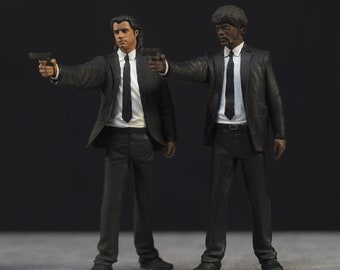 Könnte beinhalten: Zwei Actionfiguren in schwarzen Anzügen und Krawatten, eine mit einer schwarzen Afro-Perücke und die andere mit dunklen Haaren. Beide Figuren halten Pistolen und haben einen ernsten Gesichtsausdruck.