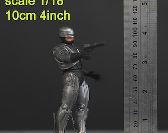 Puede incluir: Una figura de acción de un robot de color plateado con una pistola en la mano. La figura está de pie sobre una base de madera. Una regla está en el fondo, mostrando que la figura mide 10 cm o 4 pulgadas de alto. El texto "scale 1/18" es visible en la imagen.