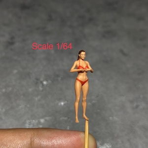 Peut inclure: Une petite figurine en bikini rouge, debout sur un cure-dent, avec le texte "Scale 1/64" au-dessus.
