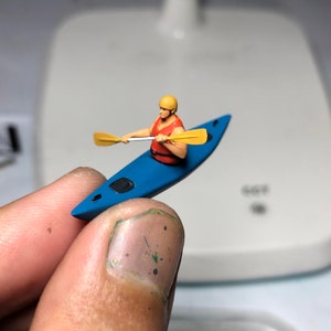 May include: A miniature figurine of a person in a red life jacket paddling a blue kayak with a yellow paddle.