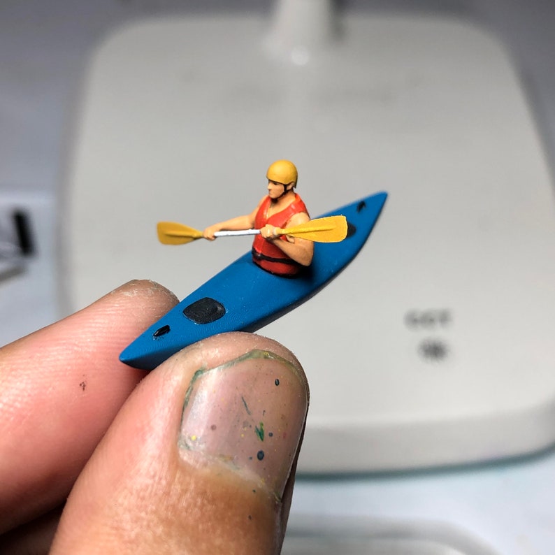 May include: A miniature figurine of a person in a red life jacket paddling a blue kayak with a yellow paddle.