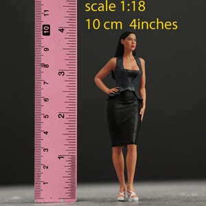 Puede incluir: Una figura femenina negra y gris que lleva un vestido negro y un chaleco gris. La figura está de pie junto a una regla rosa que mide 10 cm o 4 pulgadas. El texto "escala 1:18" y "10 cm 4 pulgadas" está impreso encima de la regla.