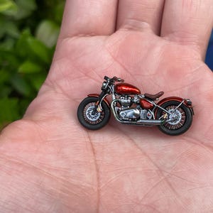Könnte beinhalten: Miniatur-Motorrad in Rot mit Chromakzenten, auf einer Handfläche liegend. Das Modell verfügt über einen detaillierten Motor, Sitz und Räder. Der Motorradkörper ist tiefrot und die Reifen sind schwarz. Ein klassisches Motorradmodell.