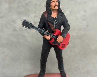 Puede incluir: Figura de un músico con un atuendo negro, tocando una guitarra eléctrica roja. La figura tiene pelo largo y oscuro y un bigote. Una cruz plateada cuelga de un collar. La guitarra tiene cuerdas y clavijas negras.