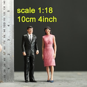 May include: Miniature figures of a man in a black suit and a woman in a pink dress. The figures are approximately 10 cm tall, with the text "scale 1:18" and "10cm 4inch" visible. The figures are standing on a gray surface.
