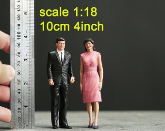 Op de afbeelding: Miniatuurfiguren van een man in een zwart pak en een vrouw in een roze jurk. De figuren zijn ongeveer 10 cm hoog, met de tekst "scale 1:18" en "10cm 4inch" zichtbaar. De figuren staan op een grijze ondergrond.