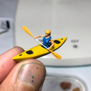 May include: A yellow kayak with a black stripe and black accents. A person in a blue life vest and yellow helmet is paddling the kayak with a yellow paddle.