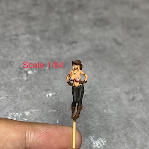Puede incluir: Una pequeña figura pintada de una mujer con un sombrero de vaquero, un top de bikini y pantalones vaqueros. La figura sostiene una pistola en una mano y un látigo en la otra. La figura está de pie sobre un palo de madera. El texto "Scale 1/64" es visible en la imagen.