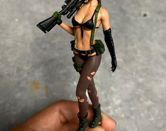 May include: A detailed figurine of a woman in a black bikini top and bottoms, green straps, and torn brown stockings, holding a black rifle. She wears black gloves and green boots. The figure is posed with the rifle over her shoulder.