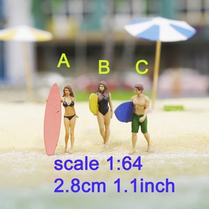 May include: Three miniature figurines of people on a beach, two women and one man, all holding surfboards. The women are wearing bikinis and the man is wearing swim trunks. The surfboards are different colors: pink, yellow, and blue. The text "scale 1:64 2.8cm 1.1 inch" is below the figurines.