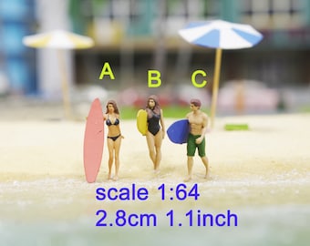 May include: Three miniature figurines of people on a beach, two women and one man, all holding surfboards. The women are wearing bikinis and the man is wearing swim trunks. The surfboards are different colors: pink, yellow, and blue. The text "scale 1:64 2.8cm 1.1 inch" is below the figurines.