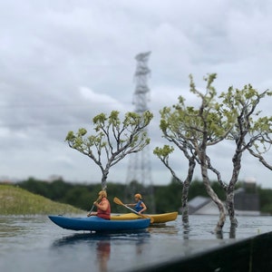 May include: A miniature scene with two figures in a blue and yellow kayak paddling on a lake. The scene is set in a miniature forest with small trees and a power line tower in the background.