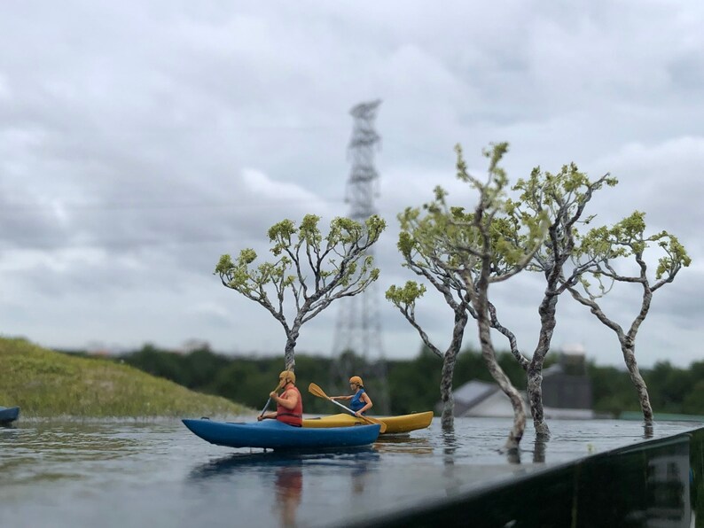 May include: A miniature scene with two figures in a blue and yellow kayak paddling on a lake. The scene is set in a miniature forest with small trees and a power line tower in the background.