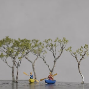 May include: Two miniature figures in kayaks paddling on a miniature lake surrounded by small trees. The kayaks are yellow and blue, and the figures are wearing red and blue shirts.