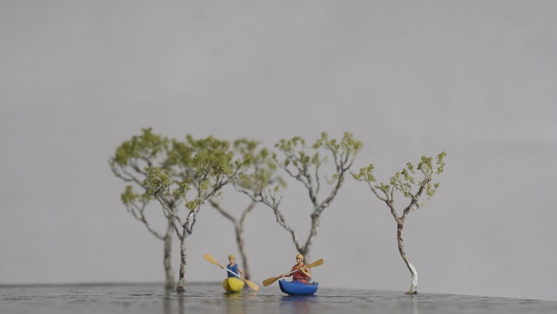 May include: Two miniature figures in kayaks paddling on a miniature lake surrounded by small trees. The kayaks are yellow and blue, and the figures are wearing red and blue shirts.