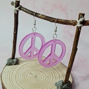 Könnte beinhalten: Ein Paar rosa Peace-Zeichen-Ohrringe, die an einem Holzständer hängen. Die Ohrringe sind aus Acryl und haben einen silbernen Haken.