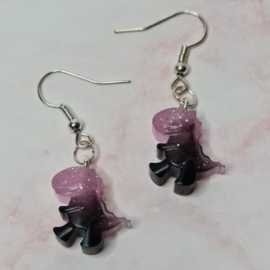 May include: A pair of silver dangle earrings with a pink and black glitter dinosaur charm.