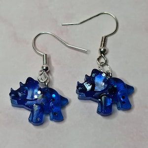 May include: A pair of blue glitter resin dinosaur earrings. The earrings are shaped like a Triceratops and have silver hooks.