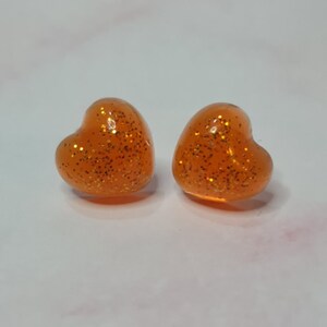 Könnte beinhalten: Ein Paar orangefarbene herzförmige Ohrringe mit goldenem Glitzer, der in das Harz eingebettet ist.