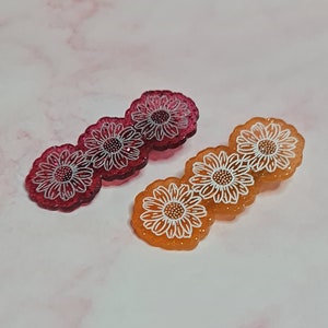 Könnte beinhalten: Zwei Sätze mit jeweils drei blütenförmigen Haarklammern. Ein Satz ist pink mit weißen Blüten, der andere ist orange mit weißen Blüten.