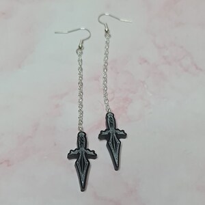 May include: A pair of silver dangle earrings with black dagger charms. The daggers have a silver outline and are detailed with a textured design.