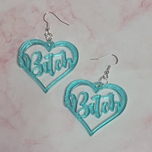 May include: A pair of turquoise blue heart-shaped earrings with the word "Bitch" written in a cursive font inside each heart.