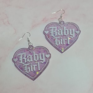 May include: A pair of purple heart-shaped earrings with white glitter and the words "Baby Girl" in white script.