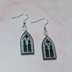 Peut inclure: Une paire de boucles d'oreilles noires avec des crochets argentés. Chaque boucle d'oreille présente un contour blanc d'une fenêtre en arc gothique.