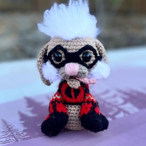 May include: A crocheted stuffed animal with a white fluffy head, black eye mask, and red and black patterned clothing. The animal has a pink nose and is sticking its tongue out.