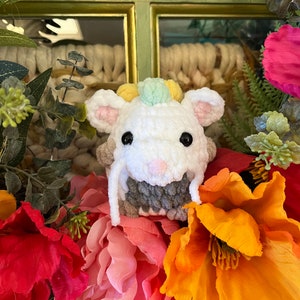 May include: A white crocheted animal with pink ears and a gray and white striped body. The animal is surrounded by pink, orange, and red flowers.
