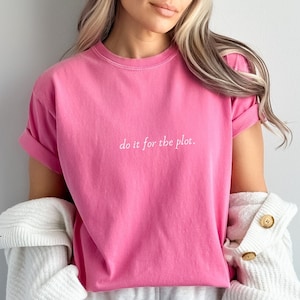 Puede incluir: Una camiseta rosa con el texto "do it for the plot." impreso en la parte delantera. La camiseta la lleva puesta una persona con el pelo largo y rubio. La persona también lleva pantalones cortos blancos y una chaqueta blanca de punto.