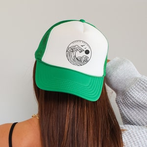 Pu&ograve; includere: Cappellino da camionista verde e bianco con una grafica circolare di un'onda, un sole e uccelli. Il cappellino ha una visiera verde e un retro in rete. Il testo "SONNIE SOCIETY" &egrave; sopra la grafica.