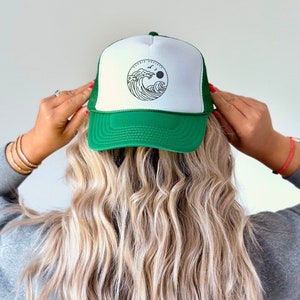 Pu&ograve; includere: Cappellino da camionista verde e bianco con un disegno circolare di un'onda, un sole e uccelli. Il cappello ha un retro in rete e una visiera curva. Il testo "Bonnie Society" &egrave; sopra il disegno dell'onda.