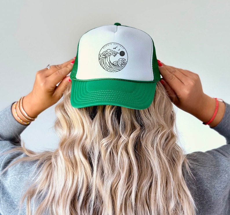 Pu&ograve; includere: Cappellino da camionista verde e bianco con un disegno circolare di un'onda, un sole e uccelli. Il cappello ha un retro in rete e una visiera curva. Il testo "Bonnie Society" &egrave; sopra il disegno dell'onda.