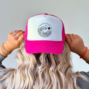 Pu&ograve; includere: Un cappellino da camionista rosa acceso e bianco con retro in rete. Il pannello frontale presenta una grafica circolare con un disegno a onde, la scritta "SUNNIE SOCIETY", un sole e uccelli.