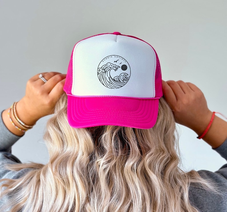 Pu&ograve; includere: Un cappellino da camionista rosa acceso e bianco con retro in rete. Il pannello frontale presenta una grafica circolare con un disegno a onde, la scritta "SUNNIE SOCIETY", un sole e uccelli.