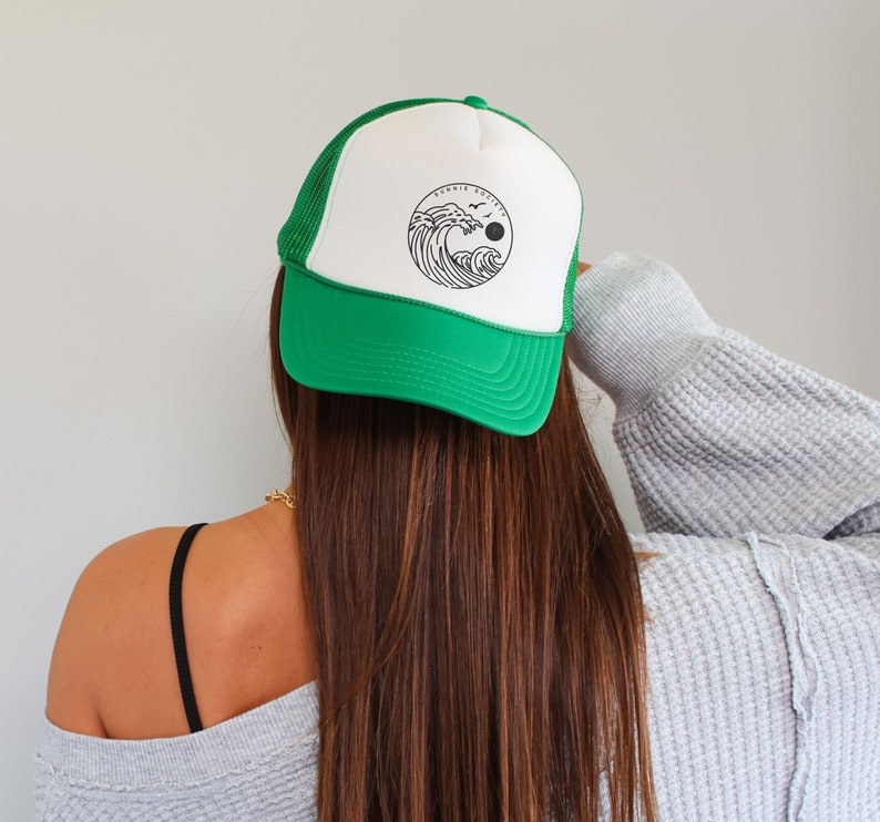 Pu&ograve; includere: Cappellino da camionista verde e bianco con una grafica circolare di un'onda, un sole e uccelli. Il cappellino ha una visiera verde e un retro in rete. Il testo "SONNIE SOCIETY" &egrave; sopra la grafica.