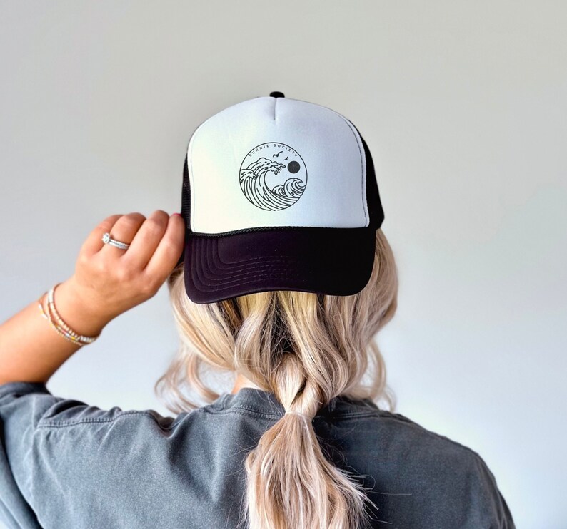 Pu&ograve; includere: Cappellino da camionista bianco e nero con un motivo circolare di onde oceaniche, un sole e uccelli. Il cappellino ha un pannello frontale bianco con la grafica e una visiera e un retro in rete neri. Il testo "SURFIN SOCIETY" &egrave; sopra la grafica.