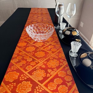 以下が含まれることがあります： 赤とオレンジの花柄のテーブルランナーが特徴のテーブルセッティング。透明なガラスのボウル、ワイングラス、装飾的なサービングピースが黒いテーブルクロスの上に配置されています。背景にはフラワーアレンジメントがあります。