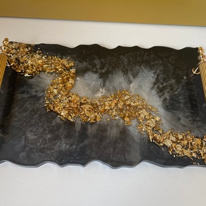 May include: Black and white resin serving tray with gold leaf accents and gold metal handles. The tray has an irregular, organic shape.