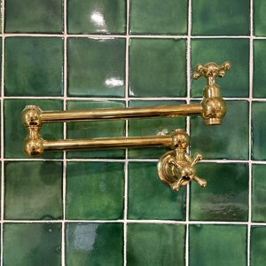 May include: A gold-toned kitchen faucet with a double-jointed arm and a cross-shaped handle. The faucet is mounted on a green tiled wall.