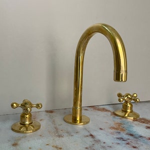 Peut inclure: Un robinet de salle de bain en or avec un bec incurvé et deux poignées en forme de croix.