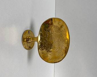 May include: A gold-toned metal wall hook with an ornate design. The hook is attached to a round base and has a curved arm.