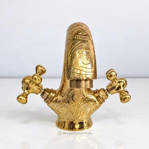 May include: A polished gold-tone metal tap featuring intricate engraved swirling patterns across the curved spout and base, with two cross-shaped handles on either side. The base includes the small text MACOPPEROFFICIAL.